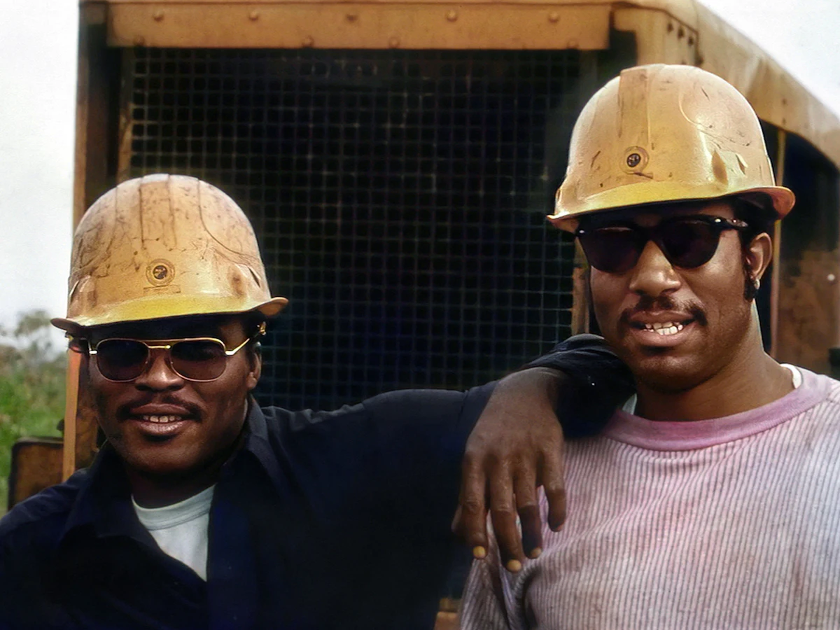 Two construction workers in hard hats on site, one with an arm around the other's shoulder.
